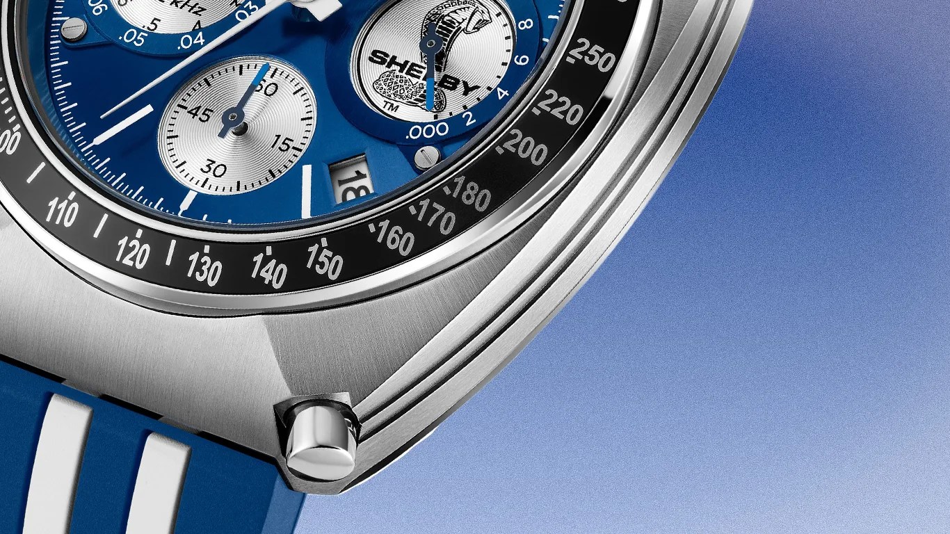 Close-up of a Bulova watch with a blue dial featuring three silver subdials, one of which has the word "SHELBY" and a cobra logo. The watch has a black tachymeter bezel with white markings and a date window showing the number 18. The case is stainless steel with a brushed finish, and the strap is blue with white stripes. A polished push button is visible on the side of the case.
