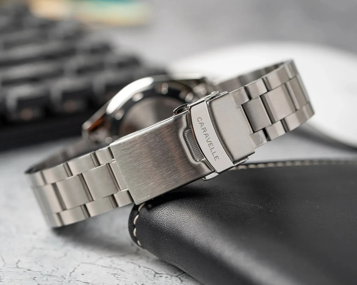 Stainless steel watch band with a brushed finish and a clasp engraved with the brand name "CARAVELLE," resting on a black leather surface with white stitching. The background is blurred, showing part of a keyboard and a watch face.