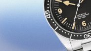 Close-up of a stainless steel Caravelle wristwatch with a black dial and bezel. The dial features beige hour markers and hands, with the text "AUTOMATIC WATER-RESISTANT 666 FEET" visible. The bezel has white numerals and dots for minute markers. The watch is set against a gradient blue background.