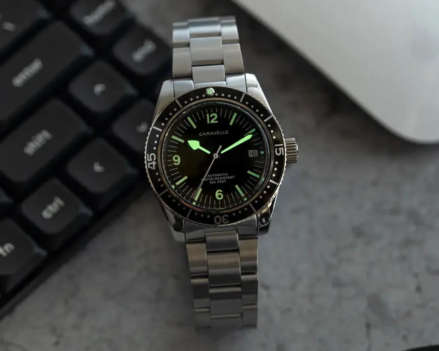 A Caravelle wristwatch with a stainless steel band and a black dial. The watch features luminous green hour markers and hands, a date window at the 3 o'clock position, and a rotating bezel with minute markings. The dial also indicates it is automatic and water-resistant to 666 feet. The watch is placed on a gray surface next to a black keyboard.