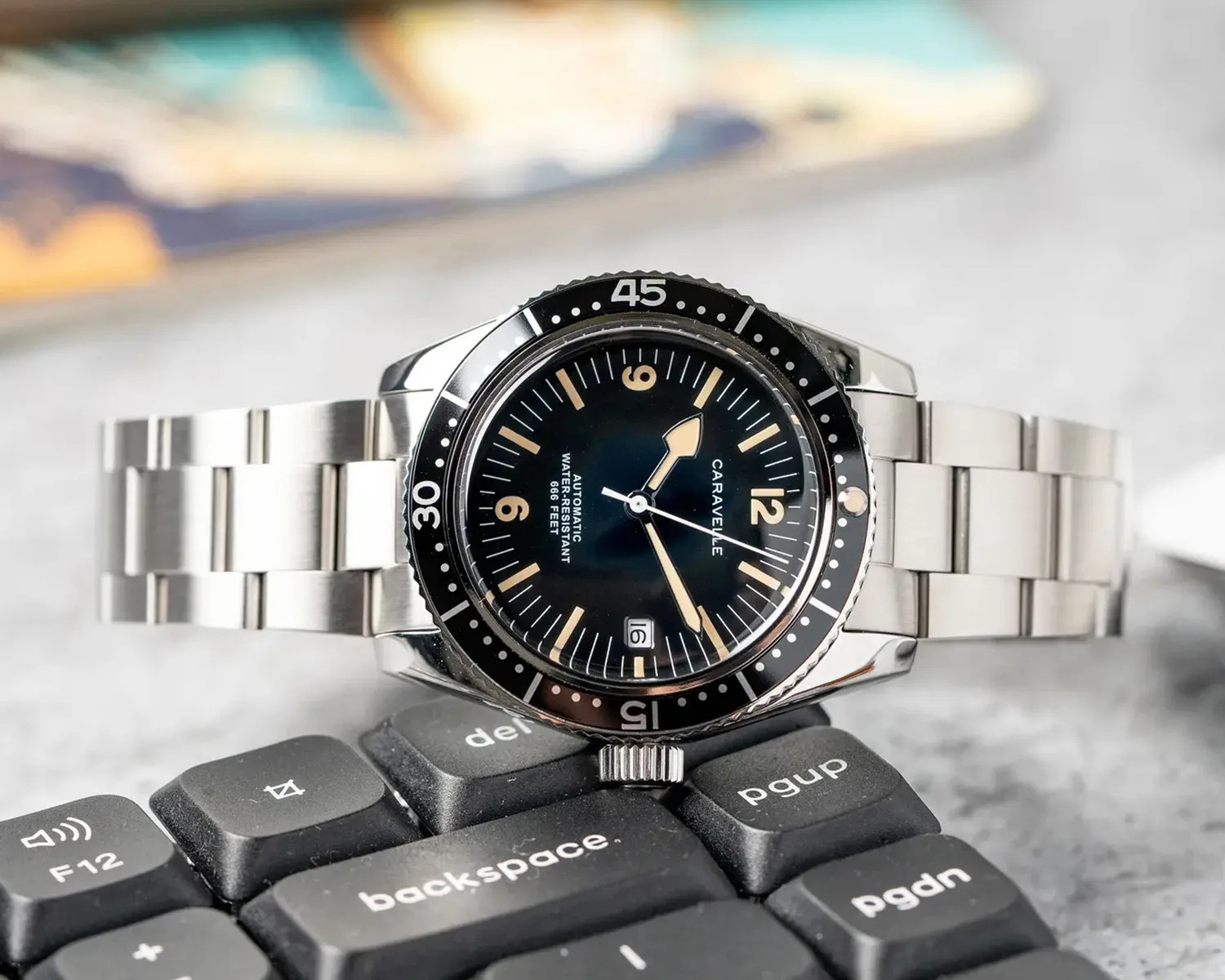 Silver metal wristwatch with a black dial and bezel, beige hour markers and hands, and a date window at the 6 o'clock position, resting on a black computer keyboard. The watch brand "Caravelle" is visible on the dial.