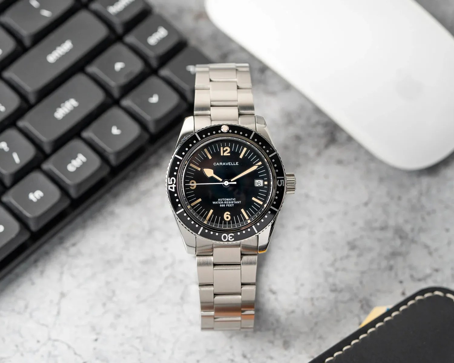 Silver metal wristwatch with a black dial and bezel, featuring beige hour markers and hands. The watch face displays the brand name "Caravelle," the date at the 3 o'clock position, and text indicating it is automatic and water-resistant to 666 feet. The watch is placed on a gray surface near a black keyboard, a white mouse, and a black leather item with white stitching.