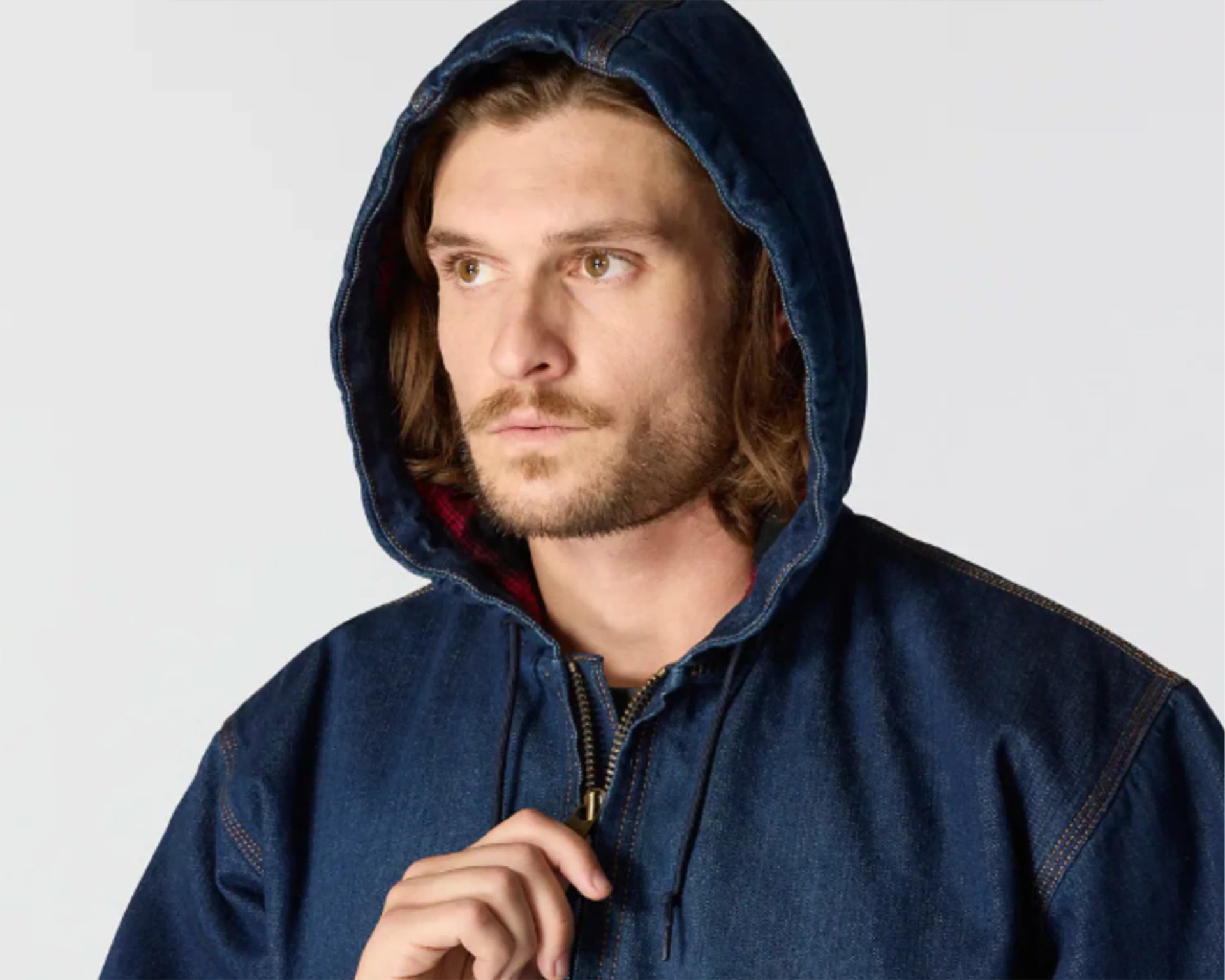 Man wearing a dark blue denim hooded jacket with a zipper, holding the zipper pull near his chest, looking slightly to the side.
