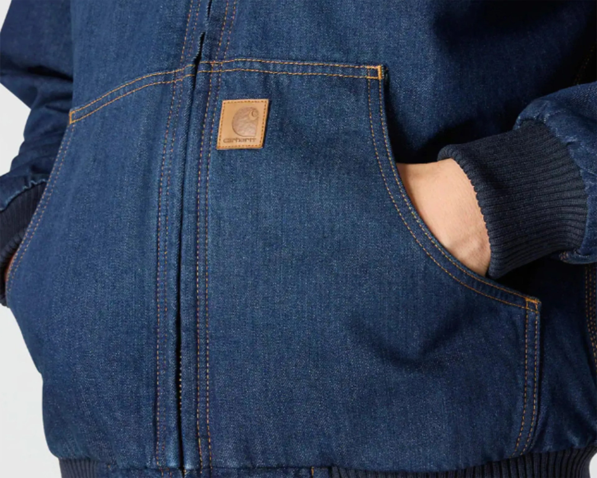 Denim jacket with orange stitching, featuring a front zipper and a small square leather patch with the Carhartt logo on the left pocket. The jacket has ribbed cuffs and waistband, and a hand is partially inserted into the right pocket.
