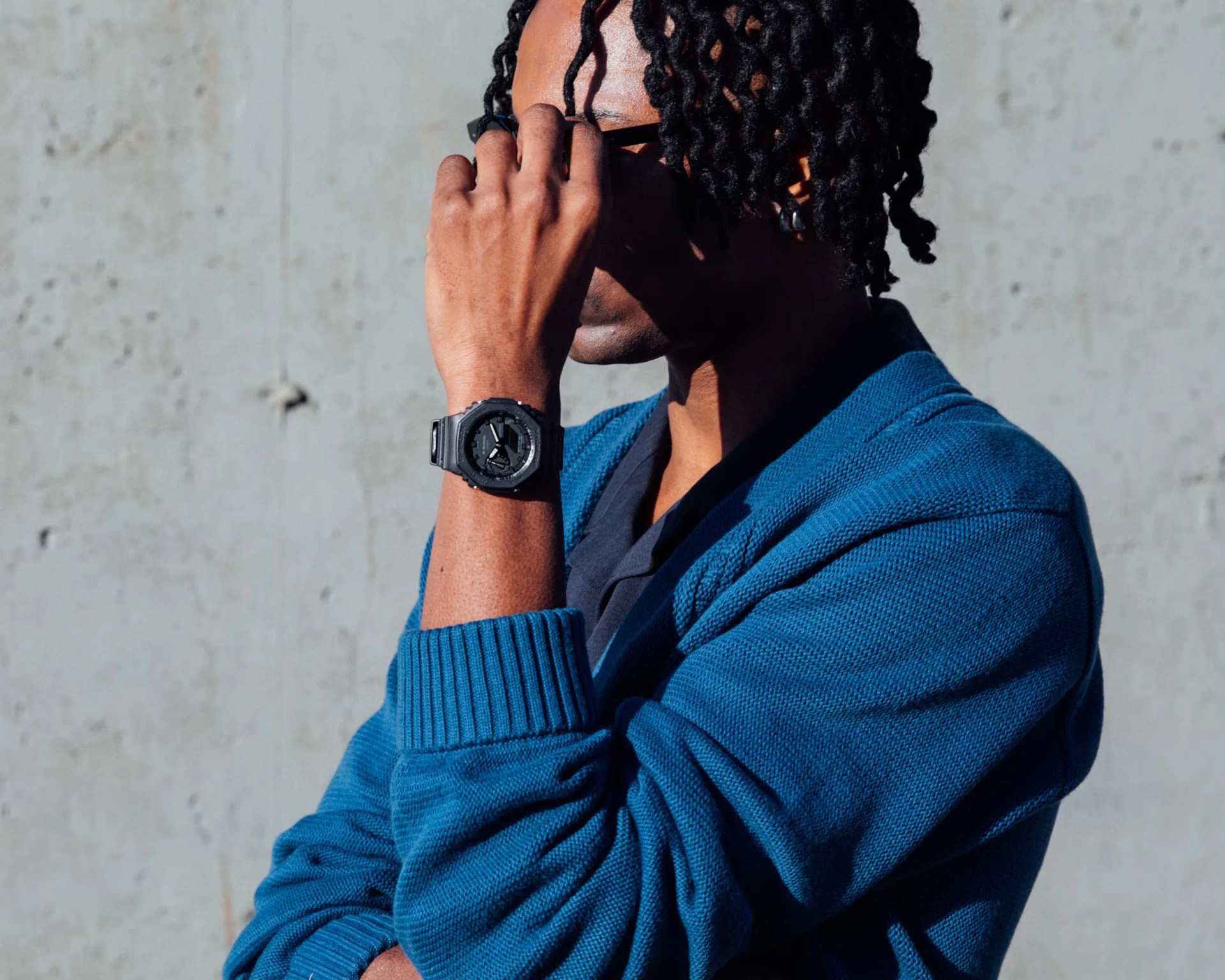 A person with dark skin and short twisted hair is wearing a blue knit sweater over a dark shirt. They are adjusting their black sunglasses with their right hand, which has a black digital wristwatch. The background is a plain, light gray concrete wall.