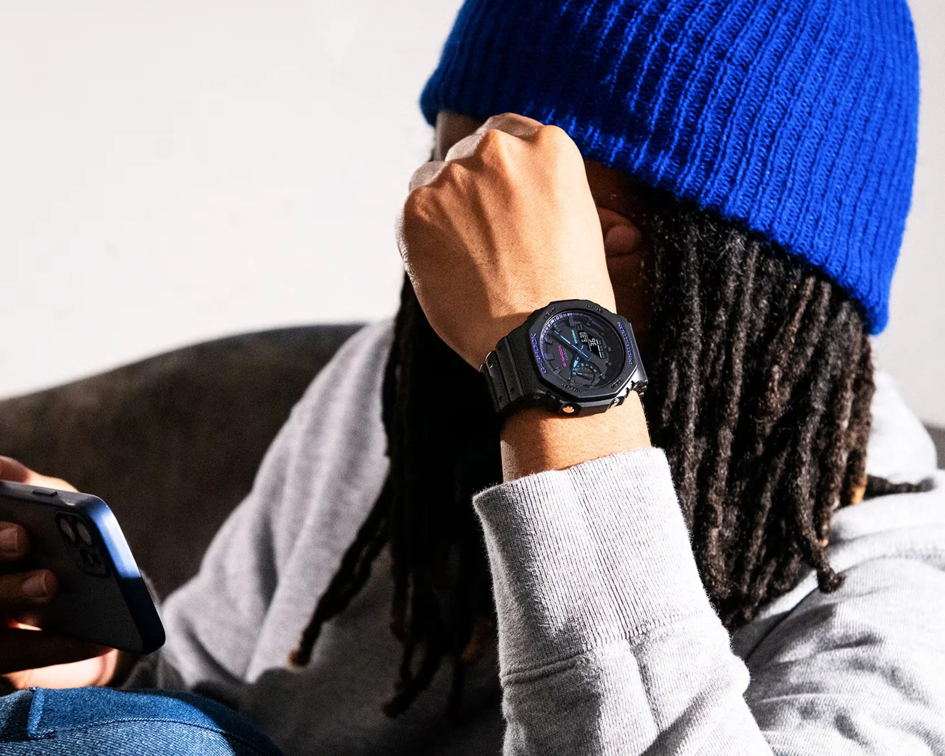 A person wearing a bright blue knit beanie and a light gray sweatshirt is holding a smartphone in one hand while the other hand, raised near their face, displays a black Casio G-Shock watch with purple and blue accents. The person has long, dark dreadlocks.