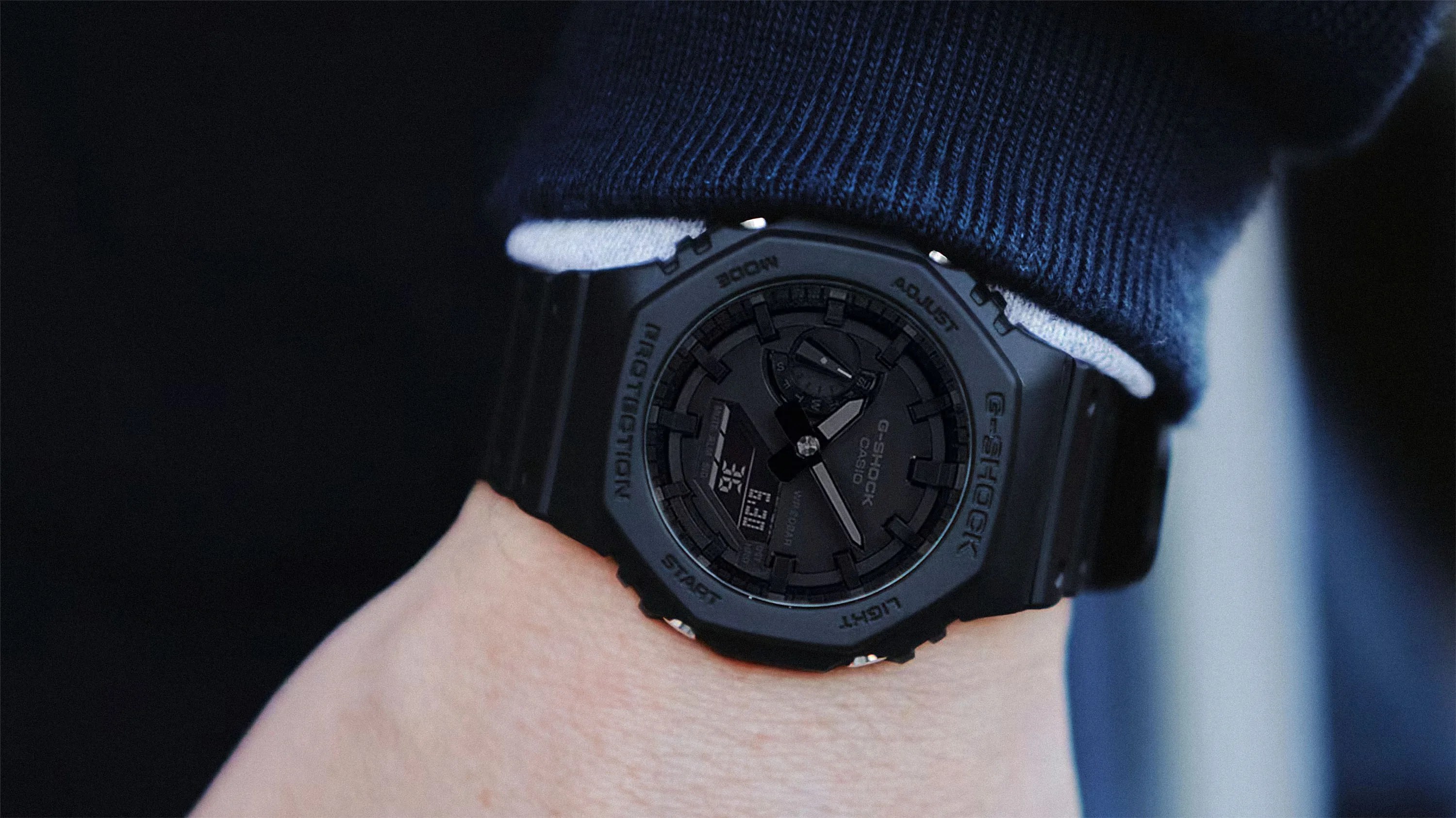Black Casio G-Shock wristwatch with a digital and analog display, worn on a wrist with a dark blue sleeve partially covering the watch. The watch has a rugged design with labels such as "PROTECTION," "MODE," "ADJUST," "START," and "LIGHT" around the bezel.