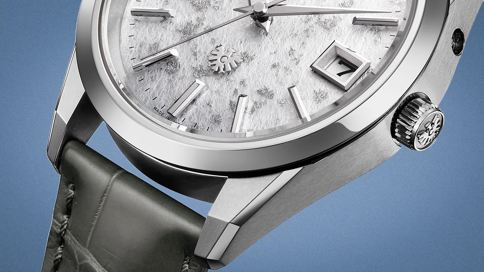 Close-up of a luxury wristwatch featuring a textured white dial with silver hour markers and hands. The watch has a date window displaying the number 7, a polished stainless steel case, and a crown with an engraved logo. The strap is dark green leather with visible stitching. The background is a solid blue color.