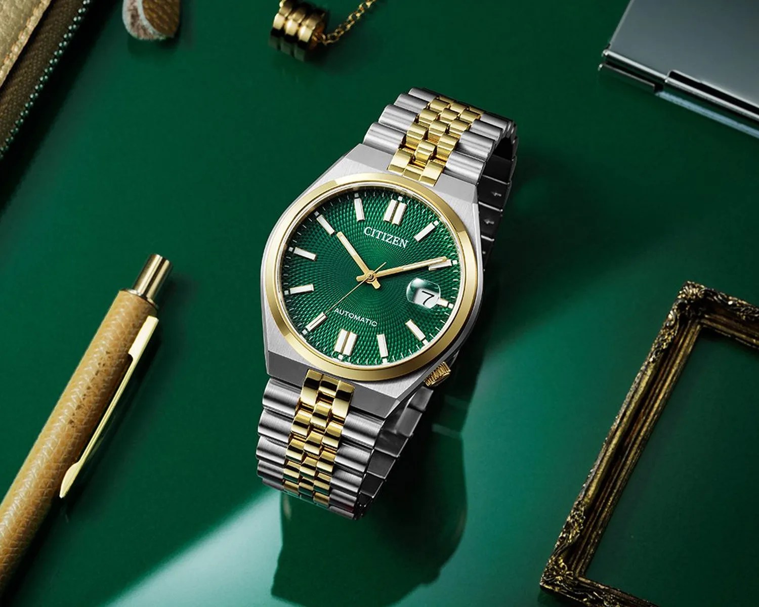 Two-tone Citizen wristwatch with a green textured dial, gold bezel, and gold and silver metal bracelet. The watch features white hour markers, gold hands, and a date window with a magnifier at the 3 o'clock position. The background is a glossy dark green surface with a gold pen, a gold chain, and a decorative gold frame partially visible.