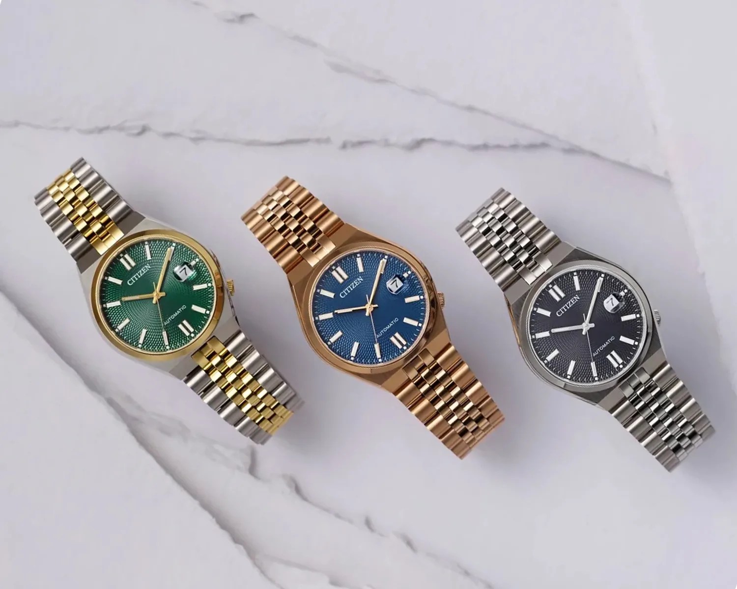 Three Citizen automatic wristwatches with metal link bracelets are displayed on a marble surface. The left watch has a green textured dial with gold and silver two-tone bracelet and gold bezel. The middle watch features a blue textured dial with a rose gold bracelet and matching bezel. The right watch has a black textured dial with a silver bracelet and bezel. All watches have white hour markers, a date window at 3 o'clock, and three hands.