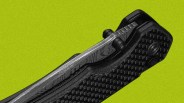 Close-up of a folding knife with a textured black handle and a partially visible blade featuring a Damascus steel pattern, set against a bright green background.