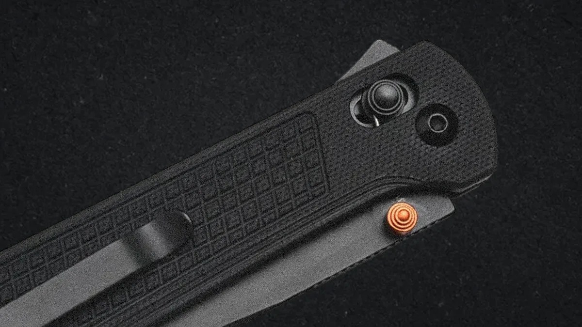 Close-up of a black folding knife handle with a textured grip, a black metal pocket clip, and an orange thumb stud on the blade. The background is a dark, textured surface.