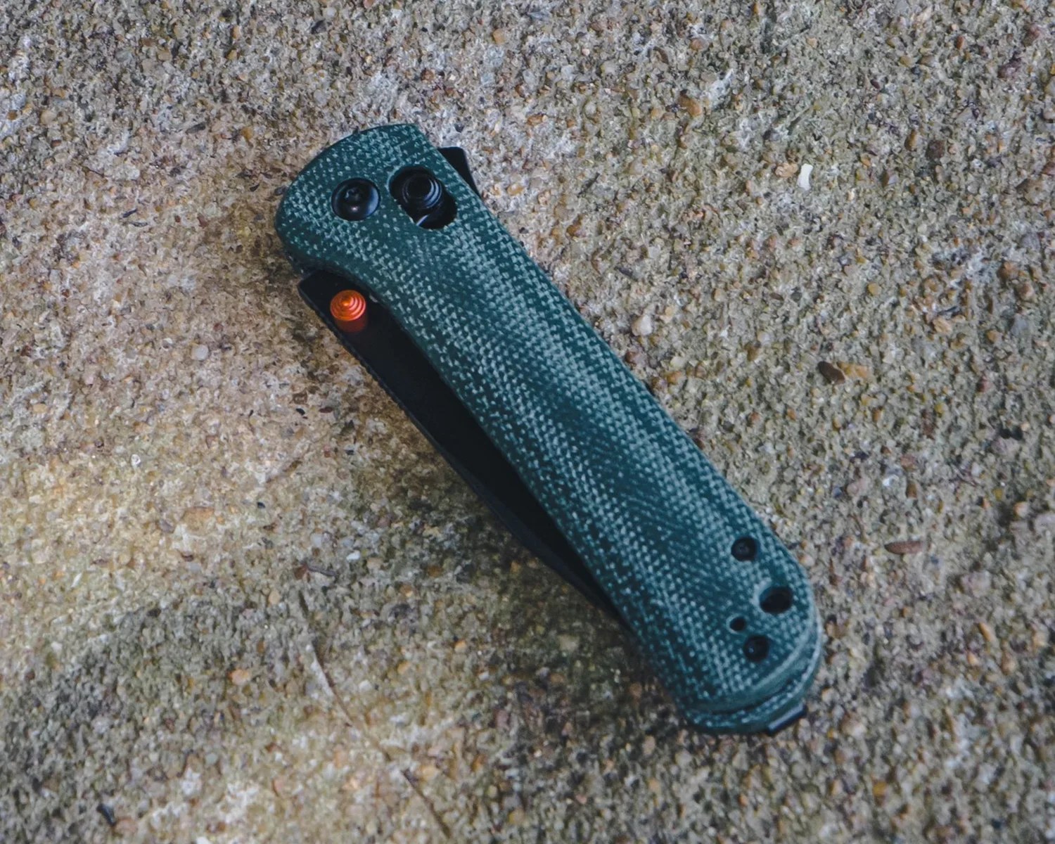A closed folding knife with a textured green handle resting on a rough, speckled concrete surface. The knife has visible screws and a small orange thumb stud near the blade pivot.