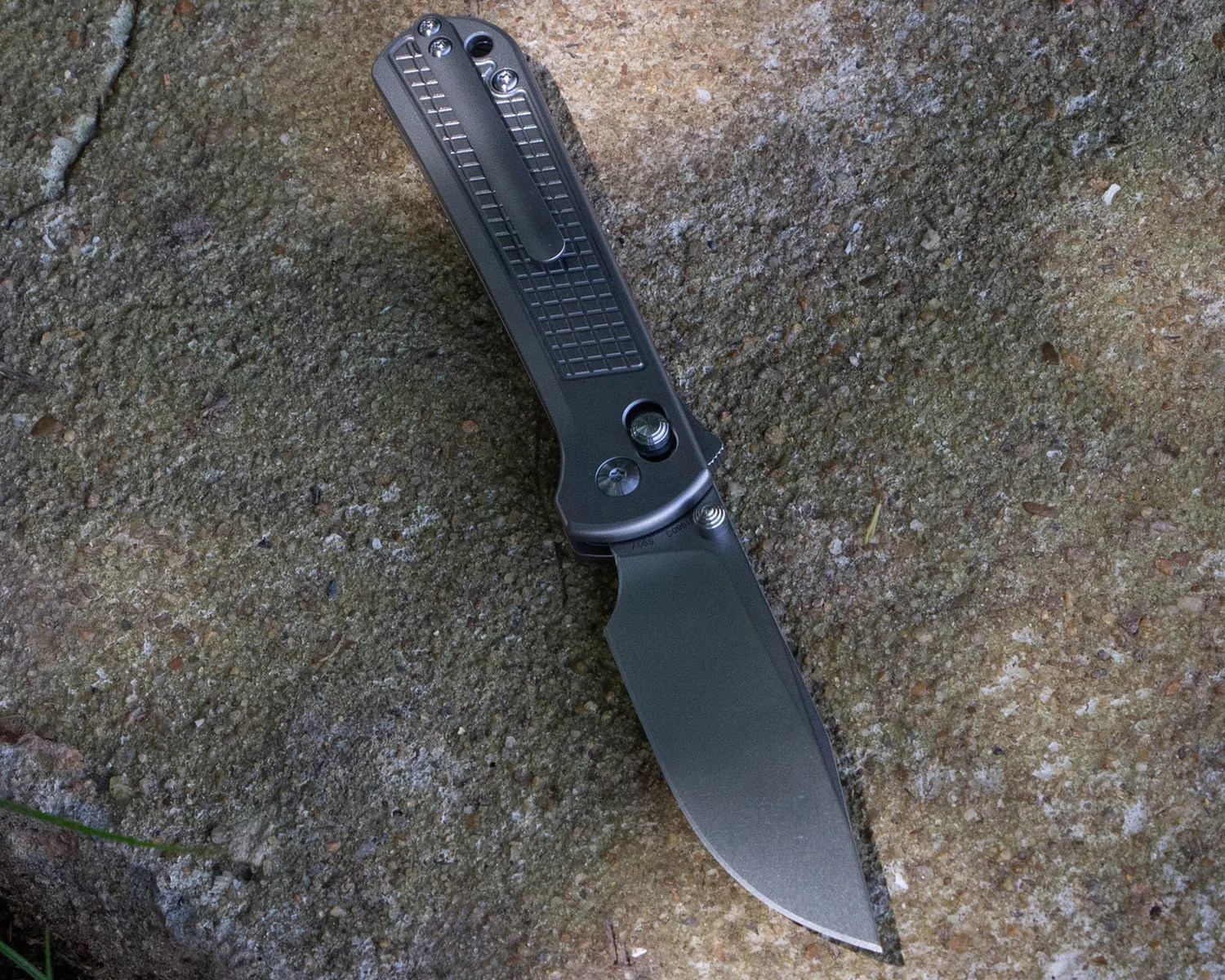 A folding knife with a dark gray handle and blade, lying open on a rough, textured stone surface. The handle features a metal clip and a textured grip area. The blade has a matte finish and a drop point shape.