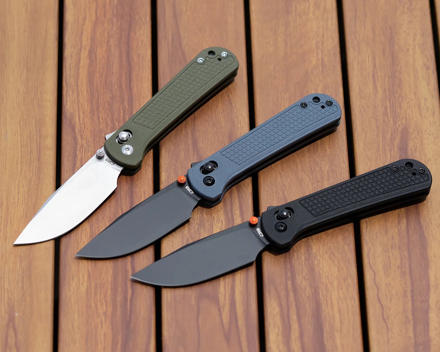 Three folding knives with textured handles are arranged on a wooden surface. The knife on the left has an olive green handle and a silver blade. The middle knife has a blue handle and a black blade with an orange thumb stud. The knife on the right has a black handle and a black blade, also featuring an orange thumb stud. All knives have a similar shape and design.