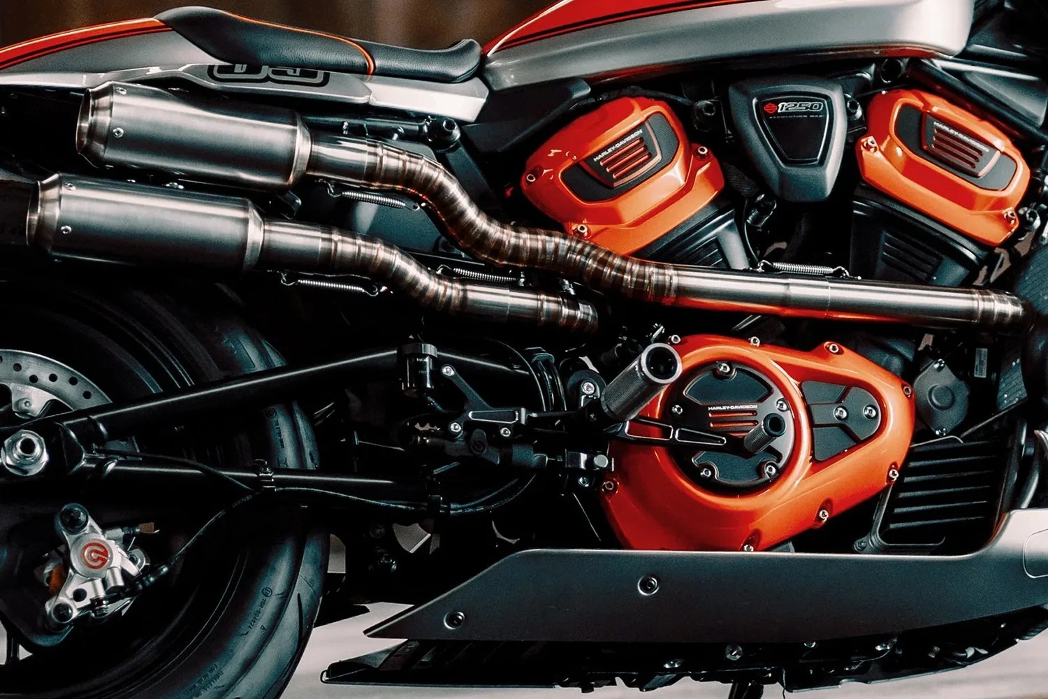 Close-up of a motorcycle engine and exhaust system featuring a black and orange color scheme. The engine has orange covers with "Harley-Davidson" branding, and the exhaust pipes are metallic with a brushed finish. The rear wheel, brake caliper, and part of the frame are also visible.