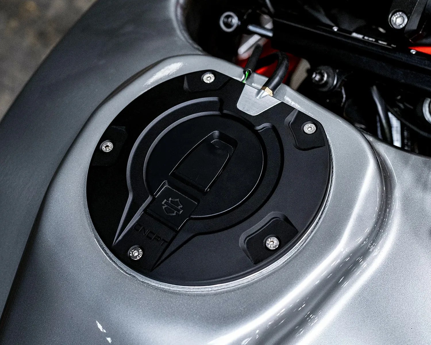 Close-up of a black motorcycle fuel tank cap mounted on a metallic silver fuel tank, featuring a locking mechanism with a Harley-Davidson logo and secured by four screws.