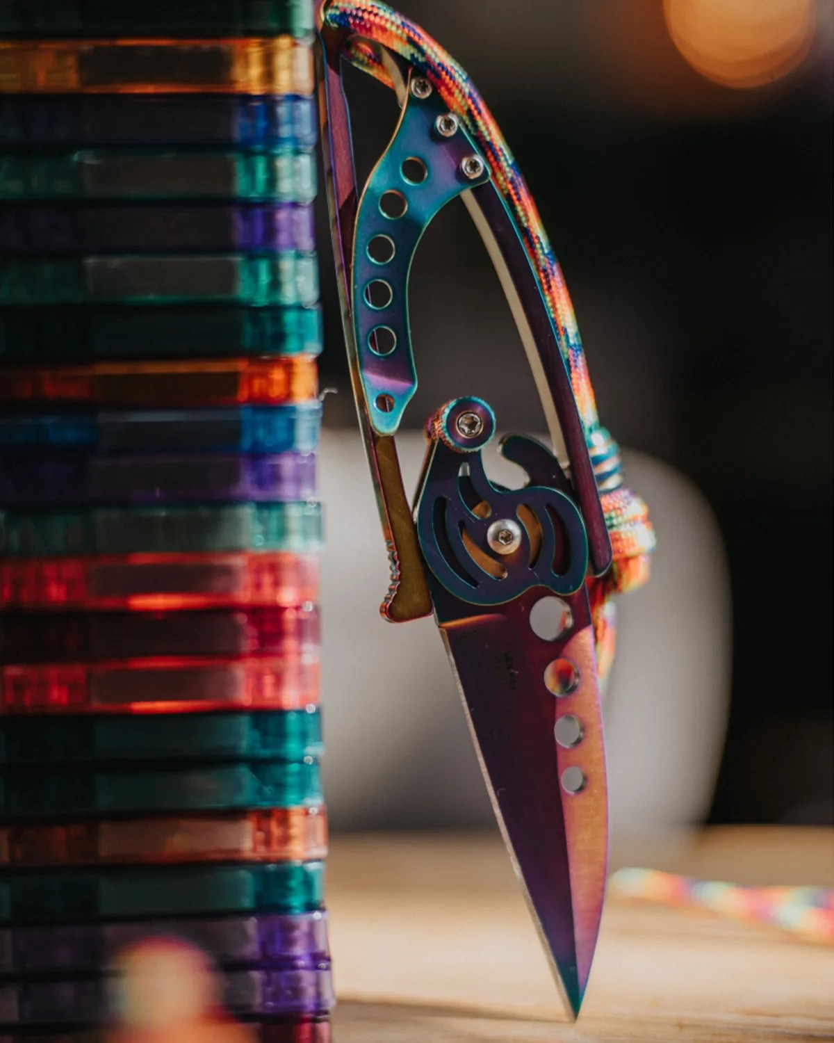 A close-up of a colorful, metallic folding knife with a rainbow finish, partially open and balanced vertically on its tip. The knife has multiple circular cutouts on the blade and handle, and a multicolored braided lanyard attached to the handle. To the left of the knife, there is a stack of translucent, multicolored plastic cases. The background is blurred with warm lighting.
