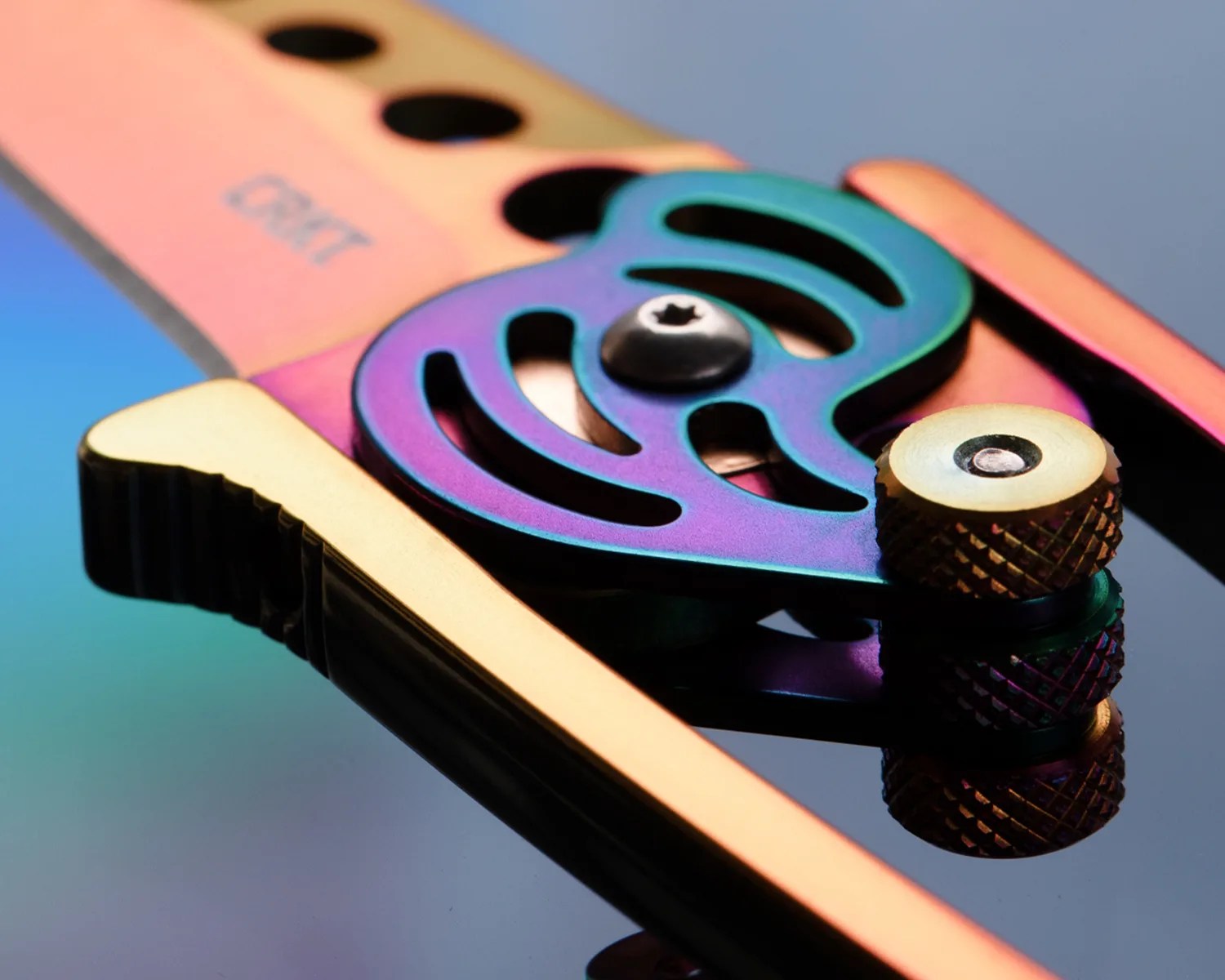 Close-up of a metallic, multicolored folding knife pivot mechanism with a textured adjustment screw and part of the blade visible, reflecting on a glossy surface.