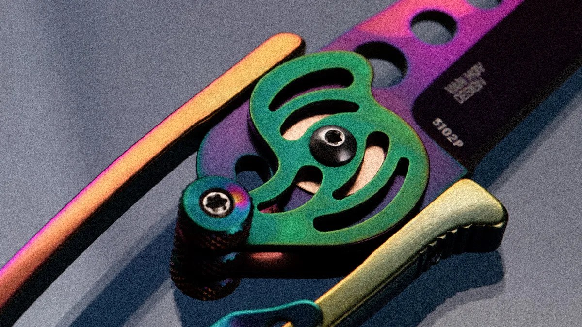 Close-up of a metallic, multicolored knife mechanism featuring a green circular component with cutouts, secured by black screws. The blade has a purple gradient with circular holes and text reading "MILITARY DESIGN" and "S102P." The handle includes a gold-toned part with a textured grip.