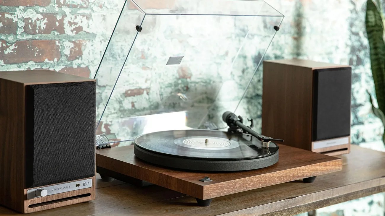 A vintage-style turntable with a wooden base and a clear dust cover is placed on a wooden surface. It has a vinyl record on the platter and a tonearm positioned over the record. On either side of the turntable, there are two matching wooden speakers with black fabric covers. The background features a textured brick wall.