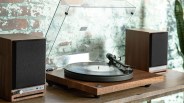 A vintage-style turntable with a wooden base and a clear dust cover is placed on a wooden surface. It has a vinyl record on the platter and a tonearm positioned over the record. On either side of the turntable, there are two matching wooden speakers with black fabric covers. The background features a textured brick wall.