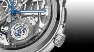 Close-up of a luxury mechanical wristwatch showing intricate gears and components. The watch has blue hands, including a second hand, and a smaller subdial with Roman numerals. The crown is visible on the right side, featuring an engraved emblem. The watch face reveals detailed craftsmanship with visible screws and jewels.