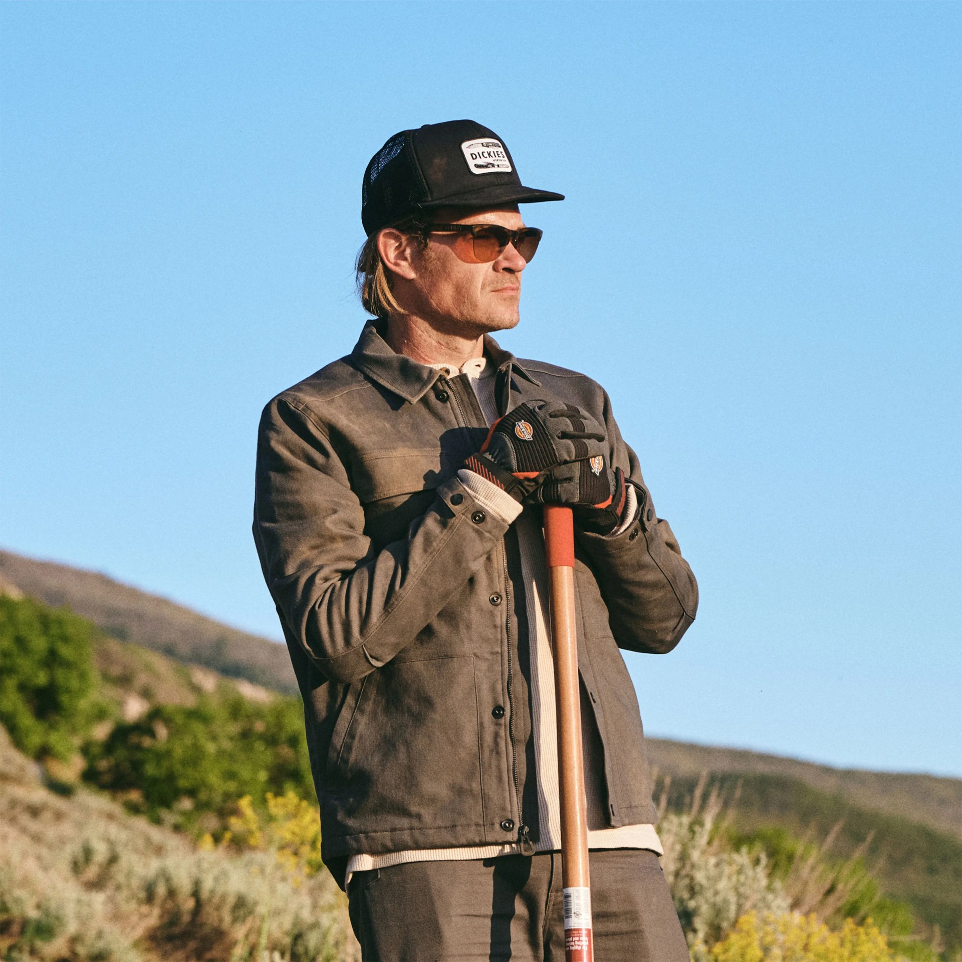 A man wearing a dark gray jacket, black gloves, sunglasses, and a black mesh cap with a "Dickies" logo, holding a wooden-handled tool, standing outdoors with a clear blue sky and hills in the background.