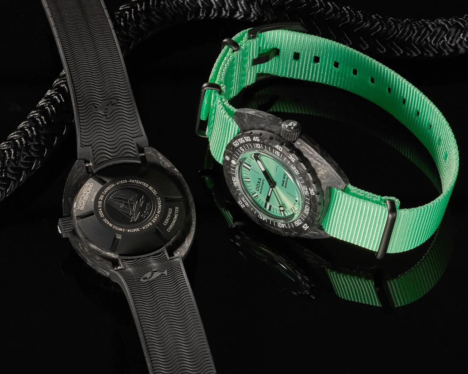 Two Doxa Sub 300 Carbon watches are shown on a reflective black surface. One watch displays the front with a mint green dial, black hands, and a black carbon bezel, paired with a bright green fabric strap. The other watch shows the back, featuring a black case back with engraved text and a sailboat logo, and a black rubber strap with a wave pattern and a small fish emblem. Both watches have a rugged, sporty design.