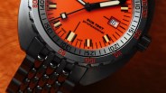 Close-up of a wristwatch with an orange dial, black and white hour and minute hands, and a black second hand. The watch face displays "SUB 750T professional" and "SWISS MADE." The date window shows the number 16. The bezel has black and red markings, and the watch has a black metal bracelet.