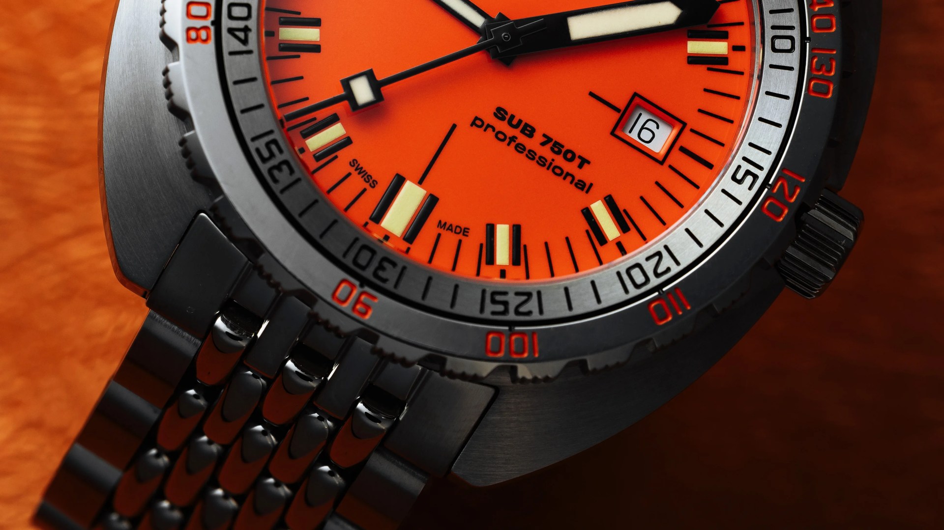 Close-up of a wristwatch with an orange dial, black and white hour and minute hands, and a black second hand. The watch face displays "SUB 750T professional" and "SWISS MADE." The date window shows the number 16. The bezel has black and red markings, and the watch has a black metal bracelet.