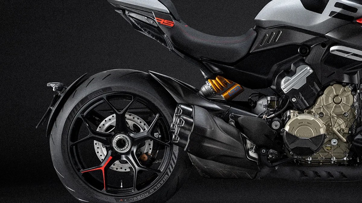 Close-up of the rear section of a motorcycle, showing a large rear tire with a black alloy wheel featuring a red accent on one spoke. The image also highlights the exhaust system with multiple outlets, a visible suspension component with a yellow spring, and part of the engine casing in a metallic finish. The seat and part of the bike's frame are also visible, with a black and gray color scheme.