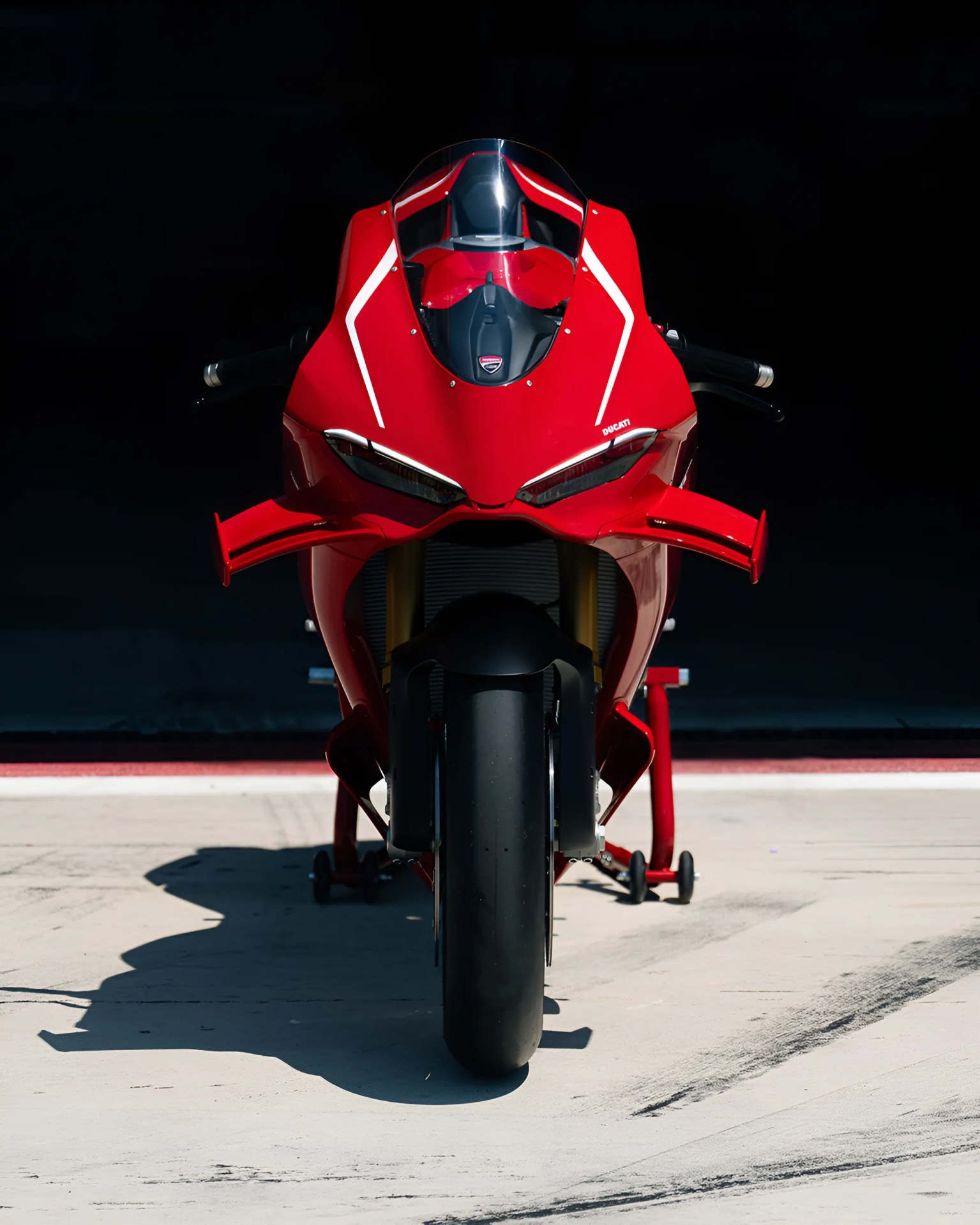 Red Ducati sport motorcycle viewed from the front, with aerodynamic winglets on both sides and a single wide front tire, positioned on a concrete surface with a dark background.