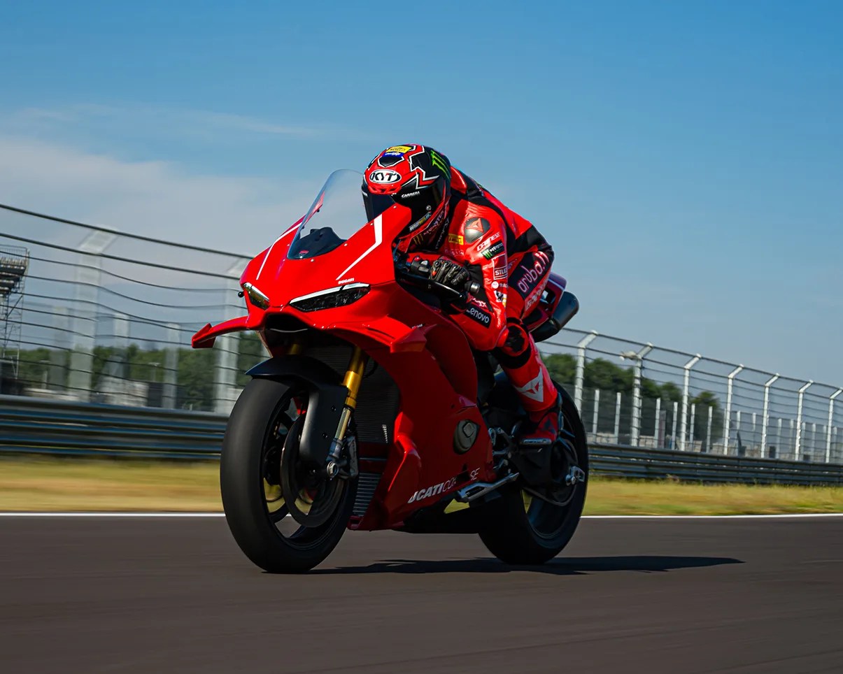 A motorcyclist in red racing gear and helmet is riding a red Ducati sportbike on a racetrack. The bike is leaning slightly forward, and the background shows a clear blue sky and a fenced area along the track.
