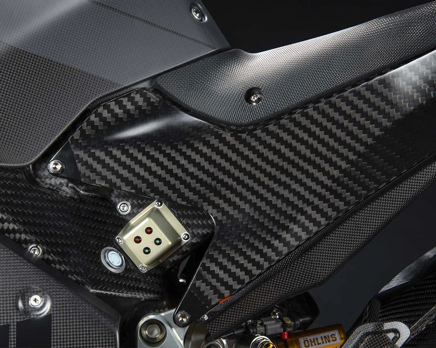 Close-up of a motorcycle frame section featuring a carbon fiber panel with a distinctive woven pattern. The panel is secured with multiple screws and includes a small metallic component with four indicator lights (two red and two green). Visible parts of the frame and surrounding components also showcase carbon fiber and textured materials, with a glimpse of an Öhlins suspension component in the lower right corner.
