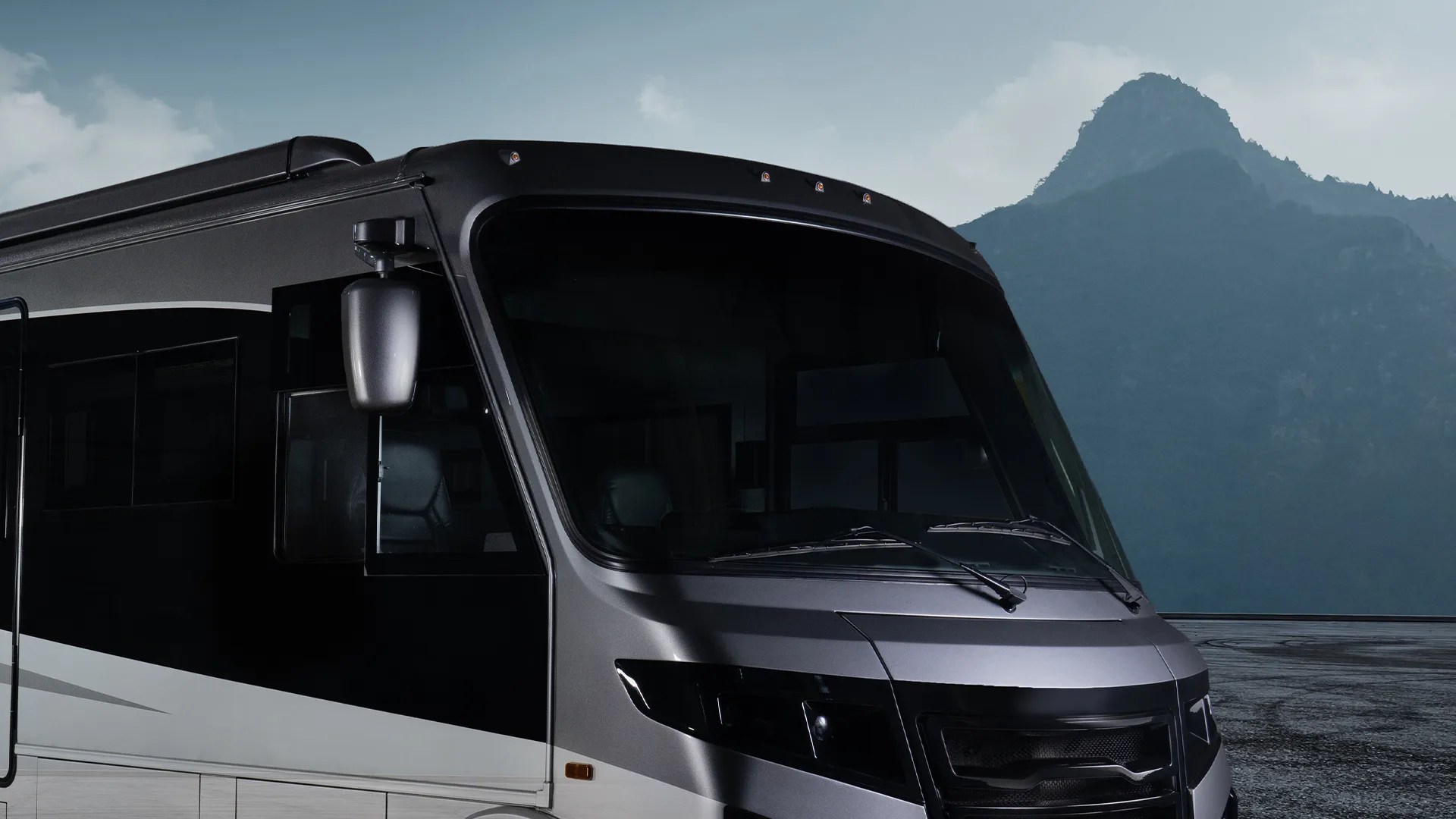 Front and side view of a modern black and gray motorhome with large tinted windows, set against a backdrop of a mountain under a cloudy sky.