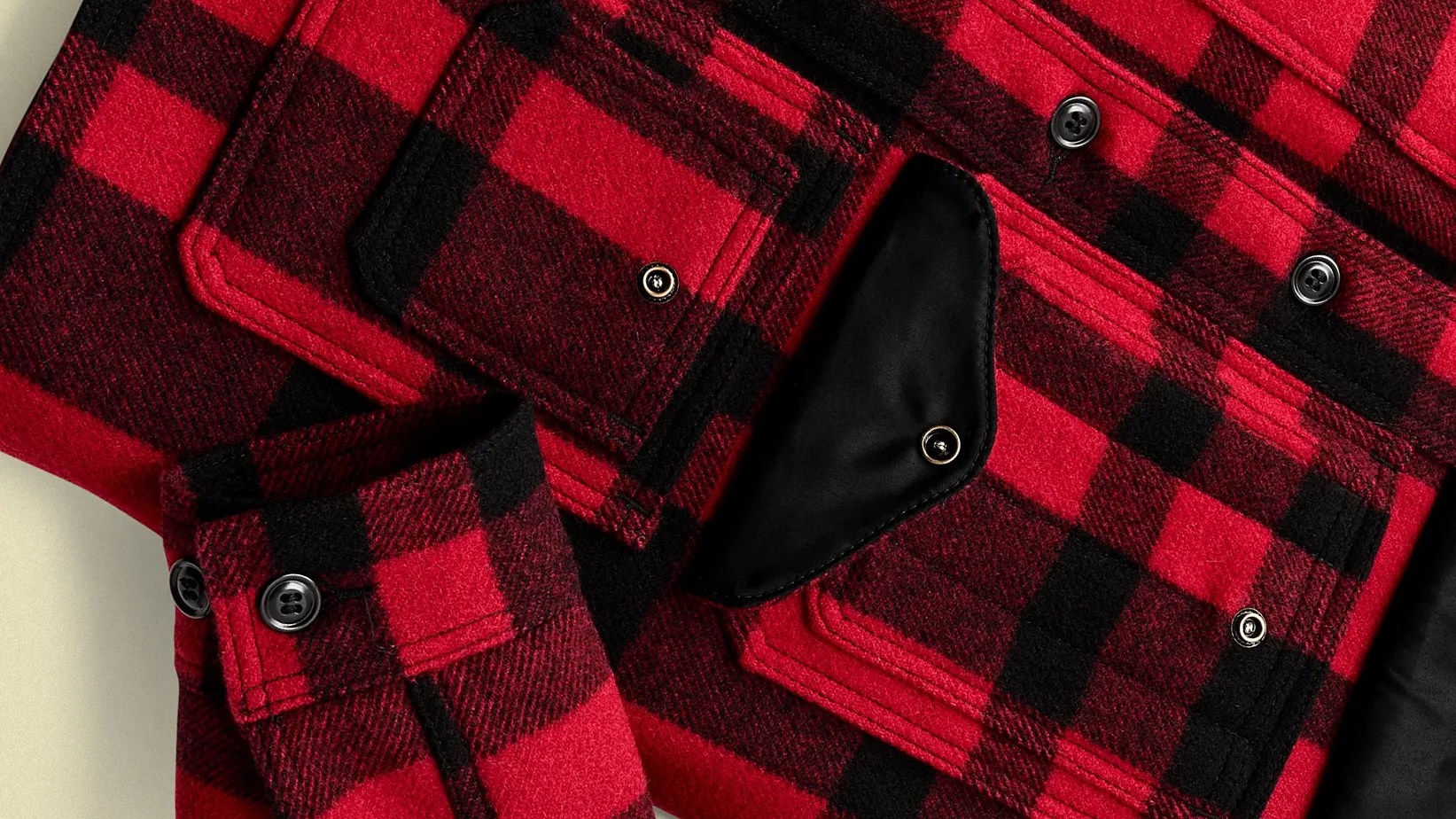 Close-up of a red and black plaid wool jacket featuring black buttons and a black leather flap pocket. The fabric has a soft, textured appearance with visible stitching along the seams and pocket edges.