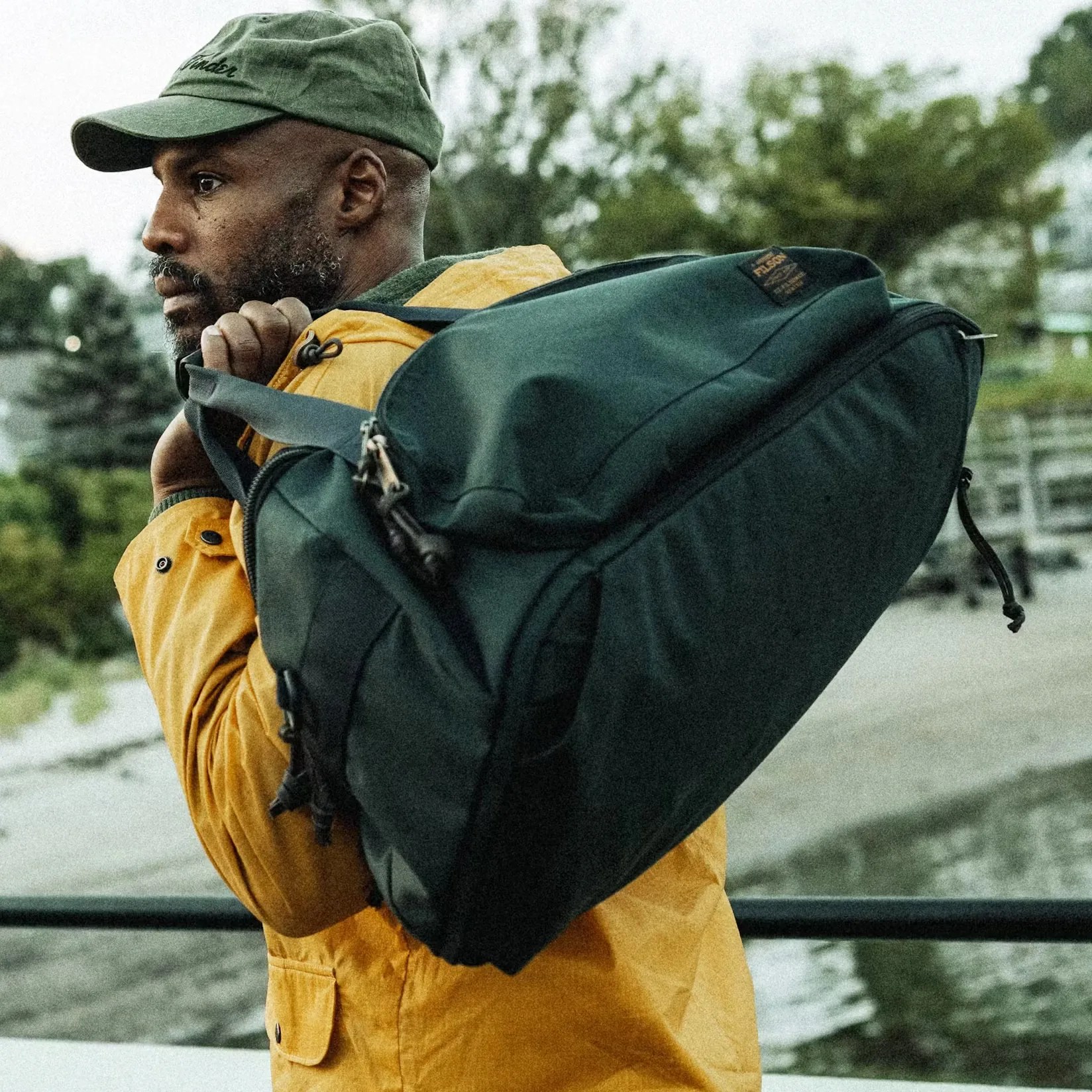 A man wearing a green cap and a yellow jacket is carrying a large dark green duffel bag over his shoulder. The background shows an outdoor setting with trees and a body of water.