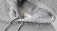 Close-up of a light gray hoodie showing the hood, drawstrings, and a fabric label inside the neckline with the text "FLINT AND TINDER." The fabric appears soft and slightly textured.