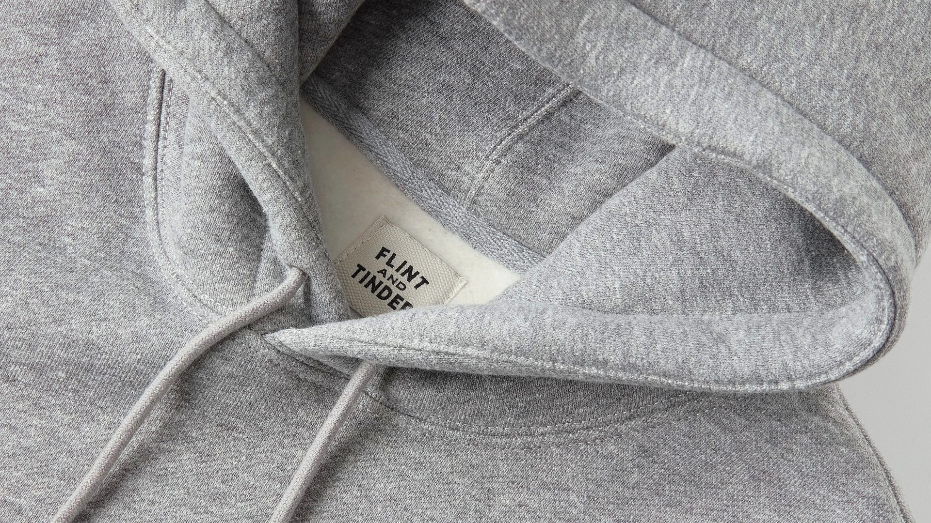 Close-up of a light gray hoodie showing the hood, drawstrings, and a fabric label inside the neckline with the text "FLINT AND TINDER." The fabric appears soft and slightly textured.