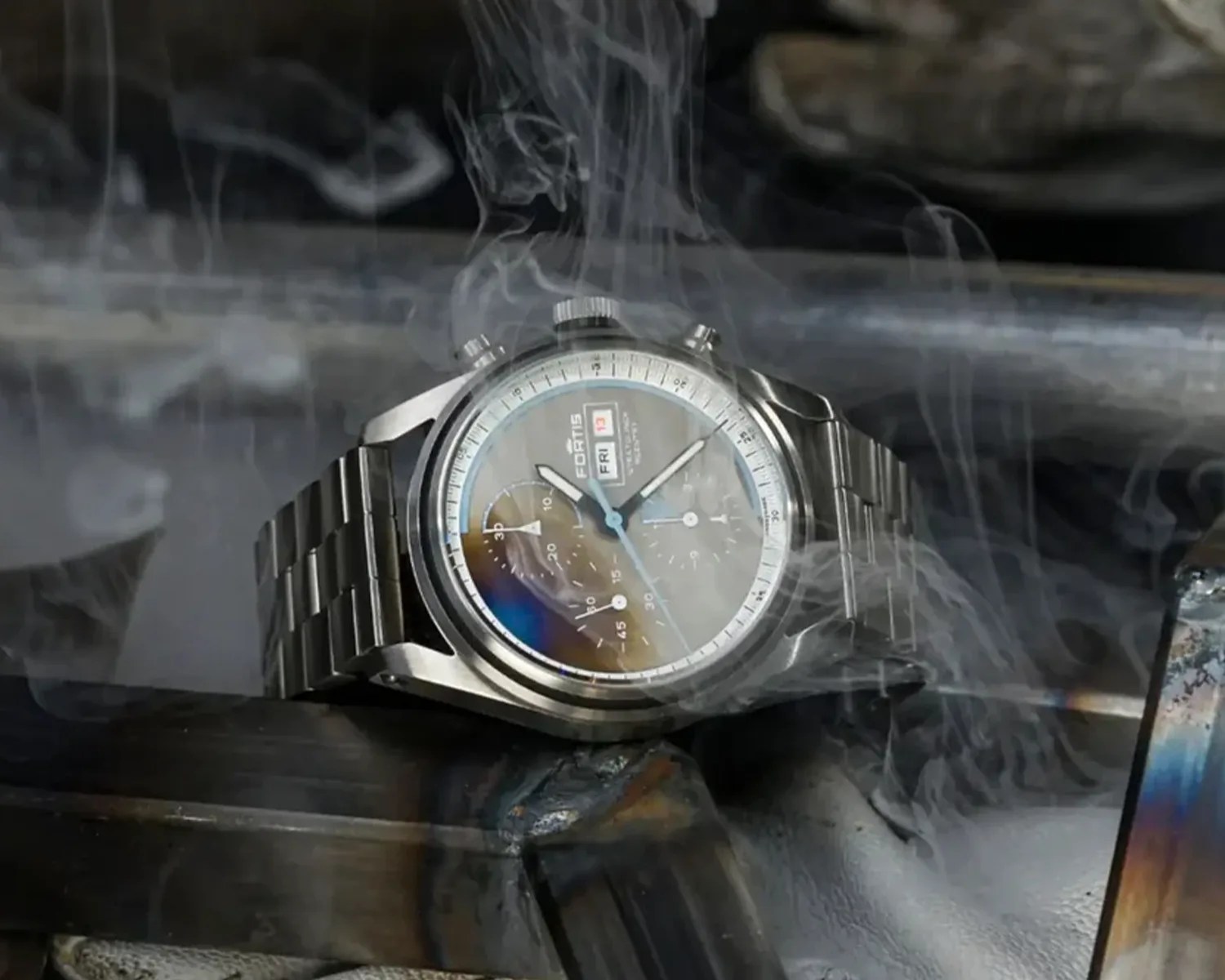 A stainless steel Fortis wristwatch with a metal link band, featuring a round dial with multiple subdials and a day-date display. The watch is placed on a dark metallic surface with smoke swirling around it.