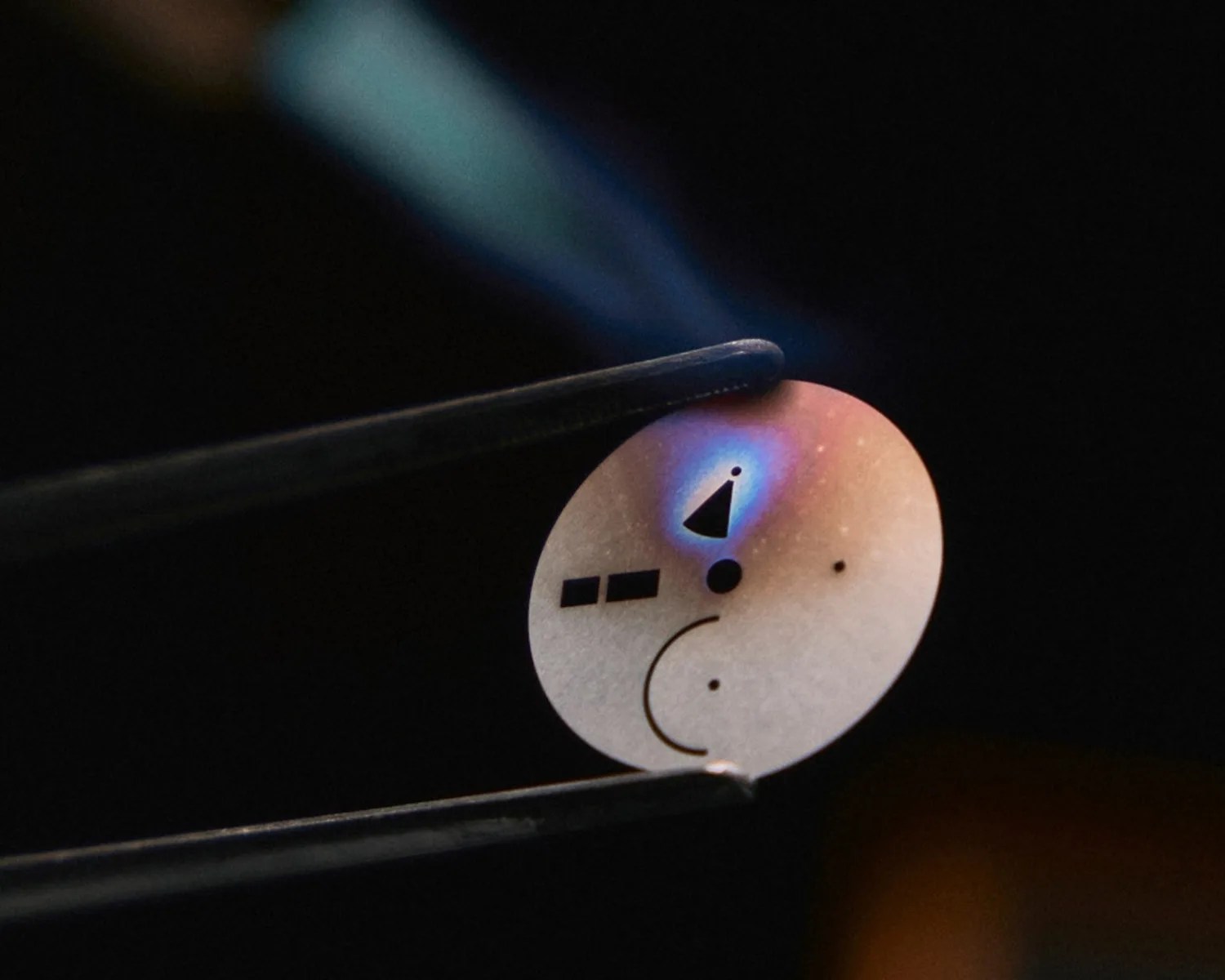 A pair of tweezers holding a watch dial with various cut-out shapes, including a triangle, circles, and rectangles. The disc is being heated, causing a blue flame and a slight discoloration around the heated area. The background is dark, highlighting the disc and the flame.