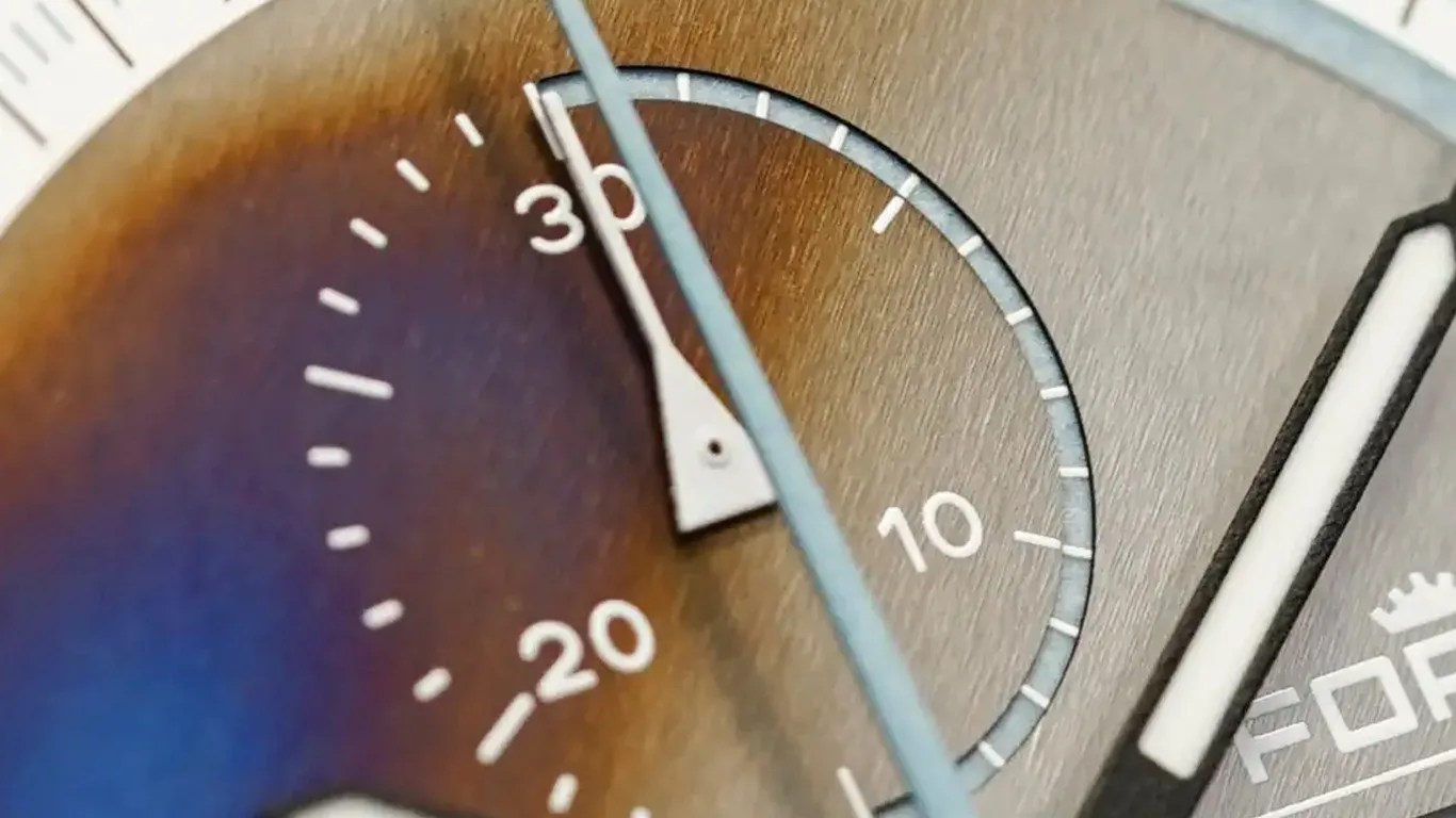 Close-up of a watch dial showing a subdial with white markings and numbers 10, 20, and 30, with a white hand pointing near 30. The background has a gradient color from blue to brown, and part of the main watch hand and the brand name "FOR" with a crown logo are partially visible. The dial has a brushed metallic texture.