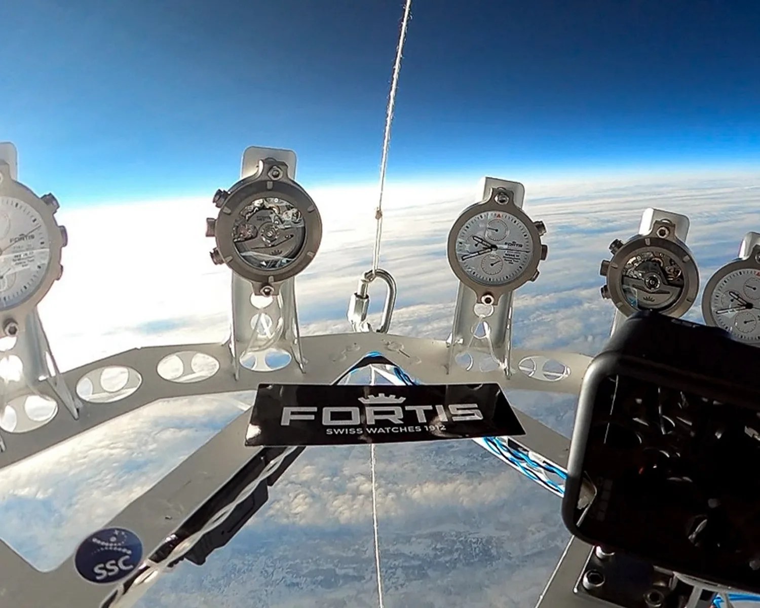A set of Fortis Swiss watches mounted on a circular metal frame, floating above the Earth with a view of the atmosphere and clouds below. The watches display both analog faces and visible mechanical movements. A black label with the Fortis logo and "Swiss Watches 1912" is attached to the frame. A camera is positioned on the right side of the frame, capturing the scene.