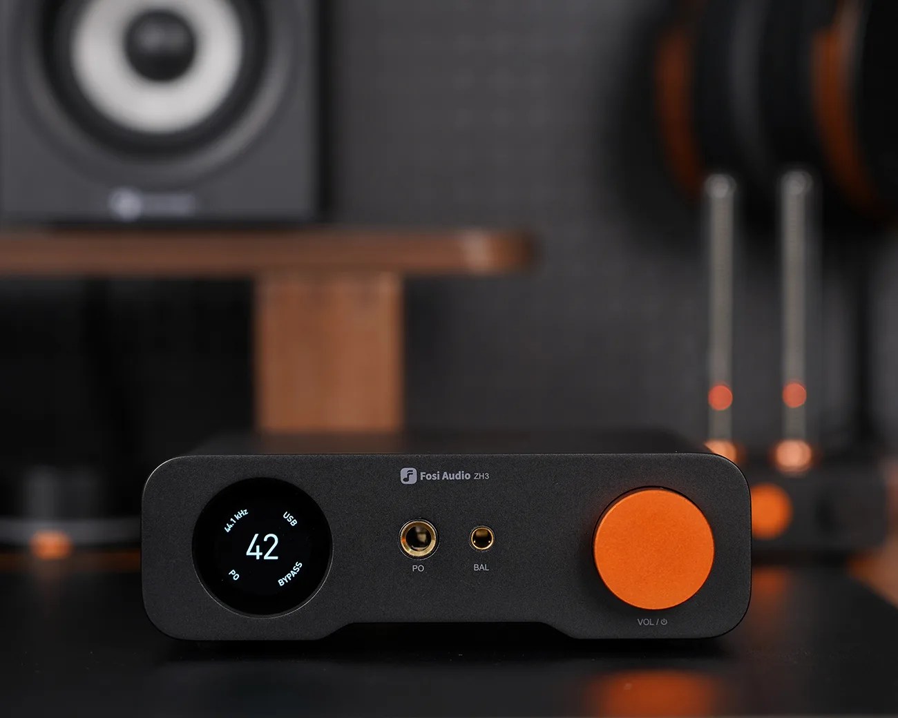 A black Fosi Audio ZH3 amplifier with a circular digital display on the left showing "42" and other text, two headphone jacks labeled "PO" and "BAL" in the center, and a large orange volume knob on the right. The background includes blurred audio equipment and a speaker.