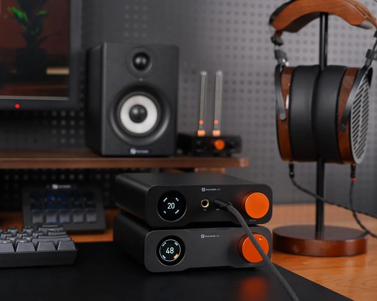 Two black Fosi Audio devices with orange volume knobs are stacked on a desk. The top device displays "20" on its circular screen, while the bottom one shows "48." A headphone cable is plugged into the top device. To the right, wooden and black headphones rest on a wooden stand. In the background, a black speaker and other audio equipment are visible.