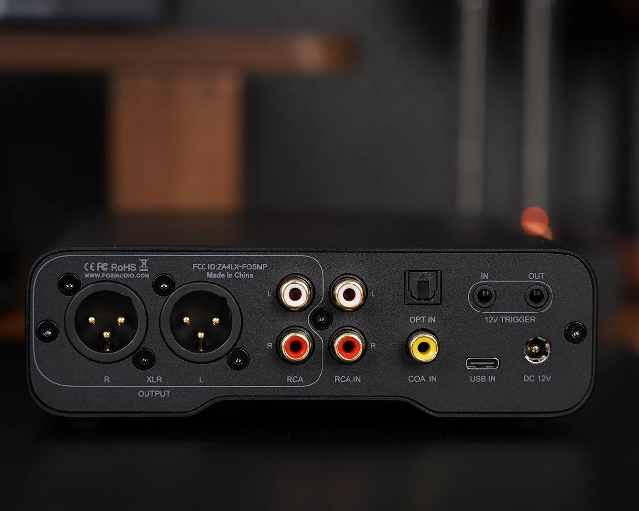 Black audio device with multiple input and output ports, including two XLR output connectors labeled "R" and "L," four RCA input connectors labeled "L" and "R," an optical input port labeled "OPT IN," a coaxial input port labeled "COA IN," a USB input port labeled "USB IN," two 12V trigger ports labeled "IN" and "OUT," and a DC 12V power input. The device has certification markings (CE, FCC, RoHS) and is marked "Made in China" with the website "FOSIAUDIO.COM.
