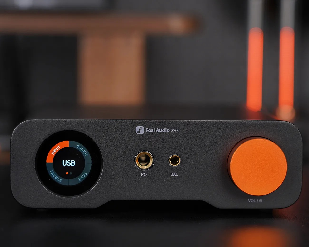 Fosi Audio ZH3 black audio amplifier with a circular display on the left showing "USB" and labeled sections for Input, Output, Treble, and Bass. It has two gold headphone jacks in the center labeled "PO" and "BAL," and a large orange volume knob on the right labeled "VOL / power.
