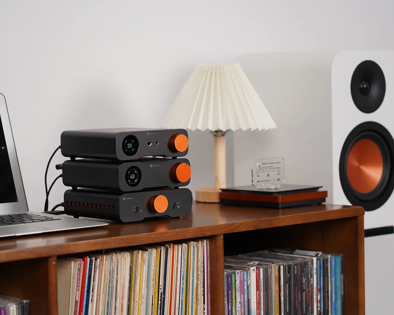 A wooden shelf holds a stack of three black audio components with orange knobs, a small lamp with a pleated white shade and wooden base, and a closed laptop. Below the shelf are vinyl records and CDs. To the right, a white speaker with an orange cone is visible.