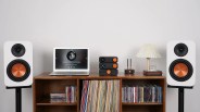A wooden shelf unit holds a collection of vinyl records and CDs. On top of the shelf, there is a silver laptop displaying a music player screen, three stacked black audio components with orange knobs, a small black device with two antennas, and a small table lamp with a pleated white shade. Two white speakers with copper-colored woofers are positioned on black stands on either side of the shelf. The background is a plain light gray wall.